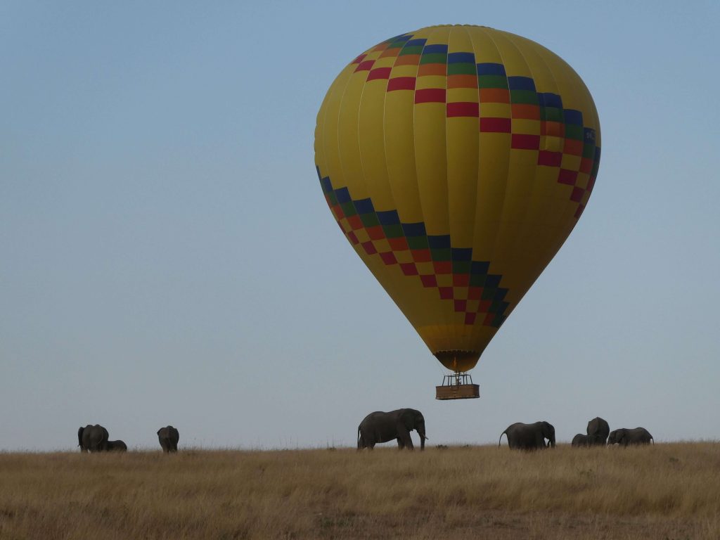 A vibrant yellow hot air balloon with a checkered pattern floating near the horizon above a herd of elephants walking across the golden savanna.